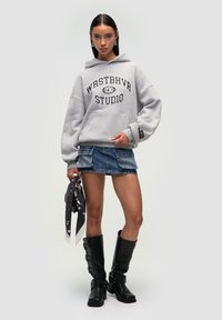 Felpa grigia con cappuccio e testo nero, gonna in denim con tasche cargo, stivali neri fino al ginocchio e una sciarpa a motivi bianco e nero.