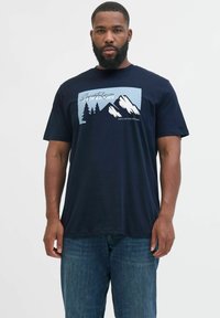 Mann mit Bart, der ein dunkelblaues T-Shirt mit einem Grafikmotiv von Bergen und Bäumen trägt, kombiniert mit blauen Jeans, vor einem einfarbigen hellen Hintergrund.