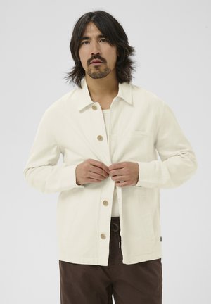 Mand med mellemlangt mørkt hår, der knappes en off-white jakke over en hvid skjorte, iført brune bukser mod en ensfarvet grå baggrund.