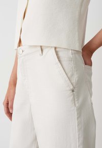 Jeans en coton beige avec une coupe décontractée, dotés d'une fermeture à bouton, de poches avant et d'une texture lisse. Coutures visibles le long des bords.