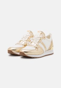 Zapatillas doradas y blancas con un acabado metálico, acentos texturizados en dorado, suela de goma y cordones. Logotipo visible en el costado.