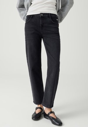 Person trägt schwarze Jeans mit geradem Bein, schwarze Ballerinas mit Schnalle, ein weißes Oberteil und einen grauen Pullover mit Händen in den Taschen.