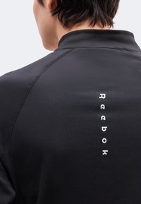 Άτομο που φοράει μαύρο μπουφάν Reebok όπως φαίνεται από πίσω, με το όνομα της μάρκας τυπωμένο κατακόρυφα με λευκό στο πίσω μέρος.