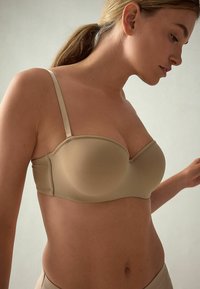 Beige strapless beha gemaakt van gladde stof, met een gevormde pasvorm en onderbroken ondersteuning, voorzien van dunne verstelbare bandjes.