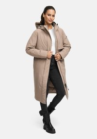 Long manteau beige matelassé avec une capuche, fermeture éclair à l'avant, poches latérales et coupe décontractée, porté par-dessus un haut blanc et un jean noir.