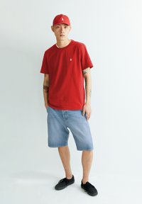 T-shirt en coton rouge avec un petit logo blanc, associée à un short en denim bleu clair. Des chaussures noires complètent la tenue décontractée.