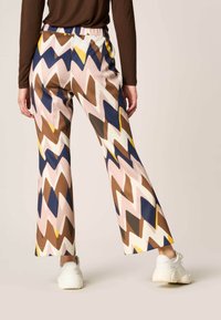 Farverige bukser med zigzag-mønster i beige, brune og blå nuancer, med en løs pasform og elastisk talje, parret med hvide sneakers.