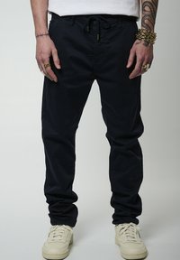 Pantaloni blu navy con vita elastica e cordoncino, caratterizzati da una vestibilità comoda e polsini. Indossati con sneakers color crema. Senza motivi visibili.