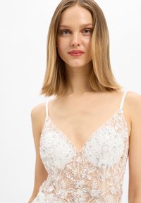 Jonge vrouw met steil blond haar, gekleed in een witte doorschijnende jurk geborduurd met bloemige kant en pailletten, tegen een effen witte achtergrond.