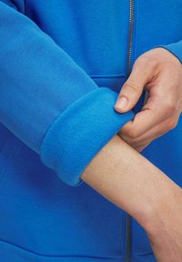 Sudadera con cremallera azul, con un puño de felpa suave, que presenta un acabado suave y una textura contrastante en el borde de la manga.