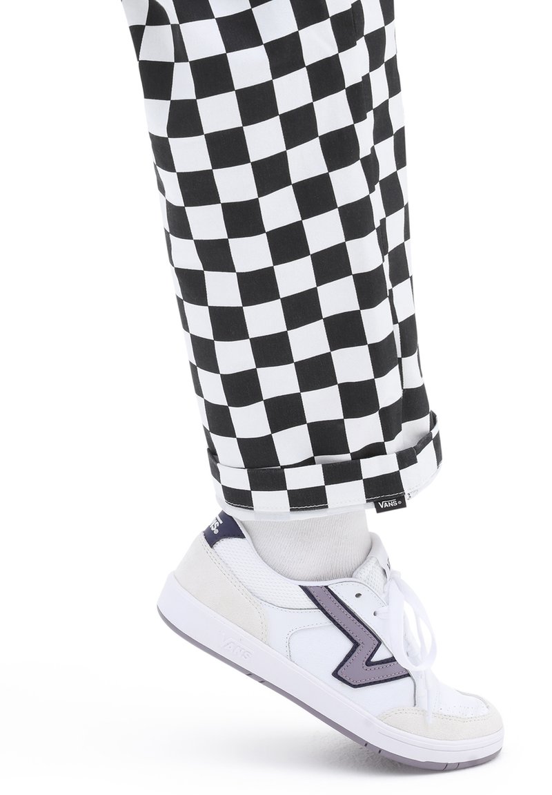 Sneakers bianchi con un accento viola, tessuto testurizzato, abbinate a pantaloni a scacchi in bianco e nero, con orlo arrotolato e etichetta con logo.