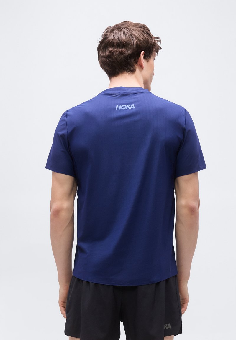 Uomo di spalle che indossa una maglietta blu HOKA a maniche corte e pantaloncini neri su uno sfondo chiaro uniforme.
