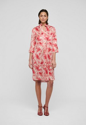 Frau steht und trägt ein knielanges, rosa-rotes Blumenkleid mit dreiviertel Ärmeln und rote Sandalen mit hohen Absätzen vor weißem Hintergrund.