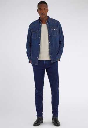Giovane uomo che indossa una camicia di denim blu scuro e jeans, t-shirt bianca sotto e stivaletti neri, in piedi con le mani in tasca.