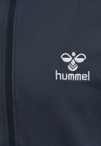 Nahaufnahme eines dunklen Stoffes mit Reißverschluss und einem weißen gestickten Bienenlogo über dem Wort „hummel“ auf der rechten Seite.