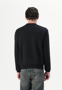 Sudadera negra con cuello redondo, tela texturizada y puños acanalados, combinada con jeans de mezclilla grises, vista desde la parte posterior.