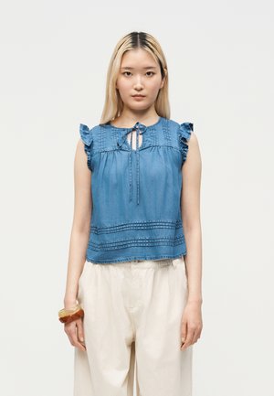Jeune femme aux cheveux blonds et raides, vêtue d'un chemisier bleu sans manches avec des volants aux épaules et d'un pantalon beige à jambes larges, debout face à l'avant.