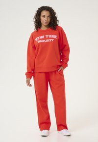 Rode fleece sweatshirt en bijpassende broek met de witte tekst "NEW YORK SIMPLICITY". De outfit heeft een relaxte pasvorm en een zachte textuur.
