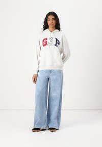 Sweat à capuche gris avec le logo "GAP" en rouge et bleu marine, coupe décontractée. Associé à un jean bleu clair à jambes larges avec un motif subtil. Chaussures noires et beiges.