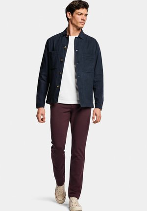Donkerblauw jack met knopen en twee voorzakken, gedragen over een wit shirt en bordeauxrode broek, gecombineerd met beige sneakers.