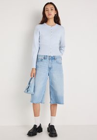 Ανοιχτό μπλε ζακέτα με κουμπιά, σε συνδυασμό με φαρδιά, κοντά τζιν σορτς σε ανοιχτό χρώμα. Μαύρα μοκασίνια και μια μικρή μπλε τσάντα συμπληρώνουν την εμφάνιση.