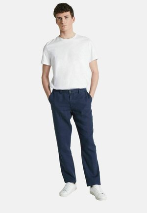 T-shirt blanc à col rond associé à un pantalon en lin bleu, avec une fermeture par bouton et des poches latérales. Porté avec des baskets blanches.