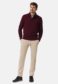 Bordeauxroter Strickpullover mit Viertel-Reißverschluss am Kragen, beige Chinos und dunkle Sportschuhe. Legere Passform, glatte Textur und neutrales Farbschema.