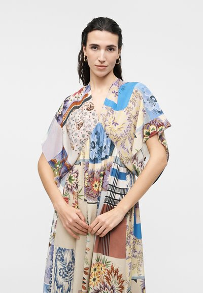 Femme aux cheveux foncés portant des boucles d'oreilles en créoles dorées et une robe patchwork colorée à motifs floraux et variés, debout devant un fond clair uni.