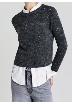 Person iført en strikket sweater med trækulspletter over en hvid skjorte med tynde sorte striber og lysegrå jeans.