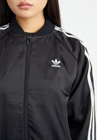 Black sporty jacket made of satin material, featuring a zip closure, white stripes on sleeves, and a small Adidas logo on the chest.