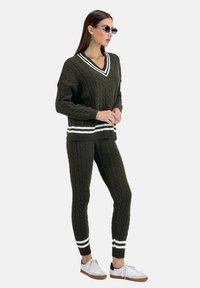 Maglione khaki e pantaloni coordinati in trama a treccia, con righe bianche su polsini e orlo. Indossato con sneakers bianche.