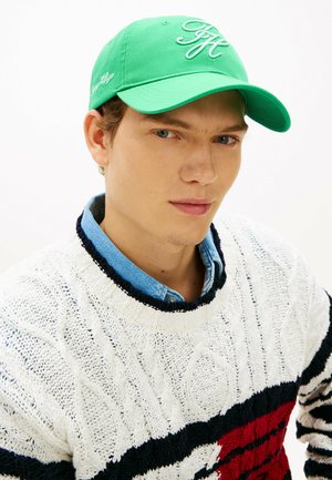 Jonge man met een groene pet, een witte kabelgebreide trui met zwarte en rode strepen, en een blauw overhemd met kraag tegen een effen witte achtergrond.