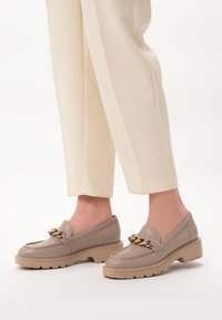 Beżowe skórzane mokasyny z grubą podeszwą, ozdobione złotym łańcuchem na wierzchu buta. Cholewka jest gładka, o zaokrąglonym kształcie palców.