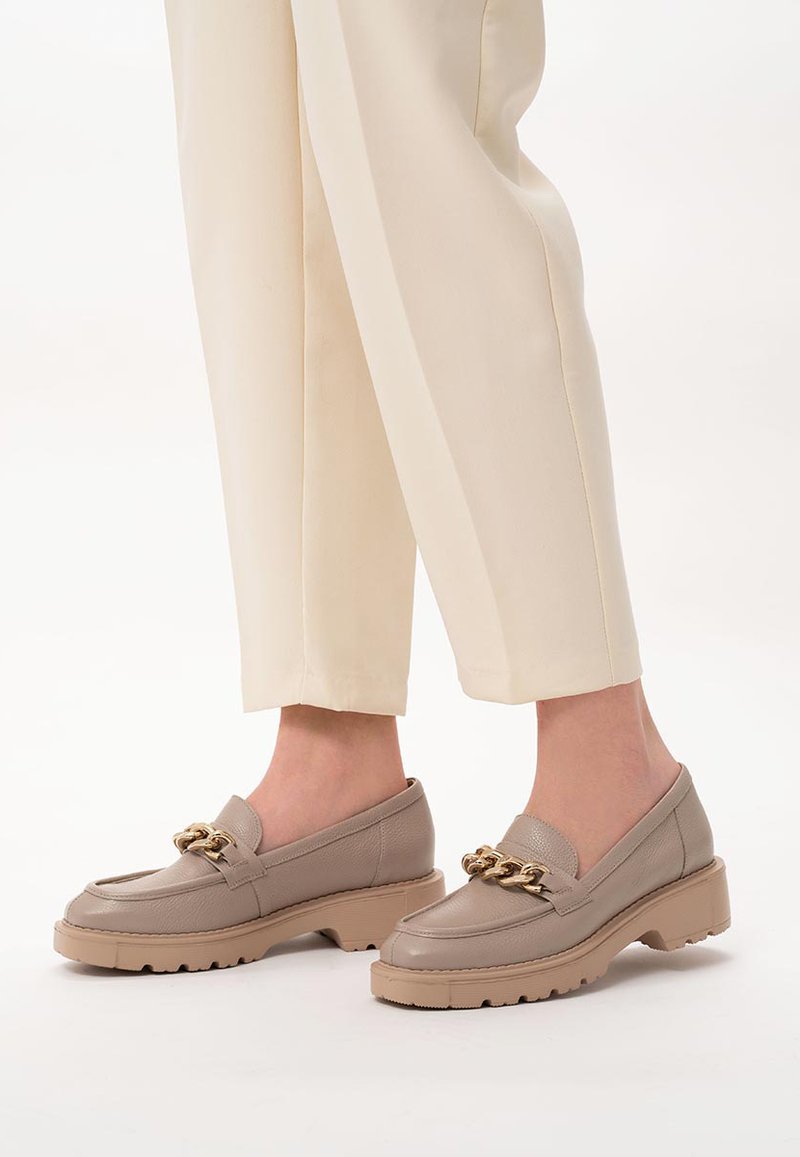 Beżowe skórzane mokasyny z grubą podeszwą, ozdobione złotym łańcuchem na wierzchu buta. Cholewka jest gładka, o zaokrąglonym kształcie palców.