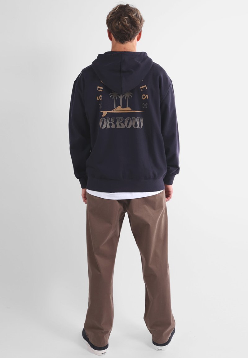 Hoodie bleu marine avec un grand graphique au dos représentant des palmiers et le mot "Oxbow" en beige, accompagné d'un pantalon marron et d'une chemise blanche en dessous.