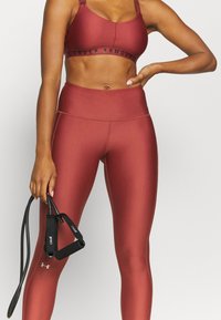 Mahonie-kleurige sport-bh met zwart logo en bijpassende hoge taille legging, beide met een gladde, rekbare textuur. Zwarte weerstandsband in de hand.