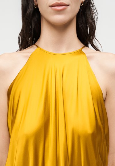 Γυναίκα που φοράει αμάνικο, μουσταρδί σατέν τοπ με λαιμόκοψη halter, με χρυσούς κρίκους στα αυτιά και χαλαρά σκούρα μαλλιά.