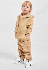 Malé dieťa so svetlými vlasmi, oblečené v béžovej mikine a zodpovedajúcich nohaviciach, stojí so zapichnutými rukami v vreckách a má na sebe biele tenisky Adidas.