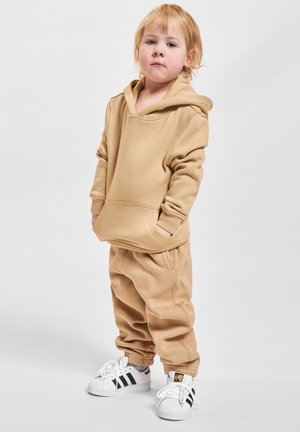 Småbarn med lyst hår iført beige hettegenser og matchende bukser, som står med hendene i lommene, iført hvite Adidas-sneakers.