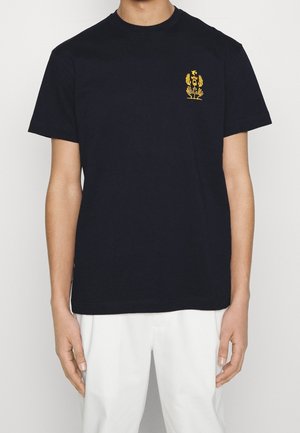 Mann trägt ein schwarzes T-Shirt mit kurzen Ärmeln und einem kleinen gelben Emblem auf der linken Brust, kombiniert mit weißen Hosen.