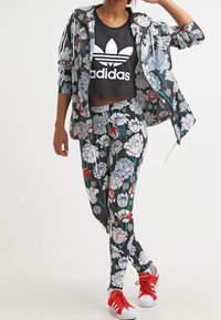 Person som bär blommönstrade leggings och jacka, svart Adidas crop top och röda Adidas sneakers, poserandes med ena handen på huvudet.