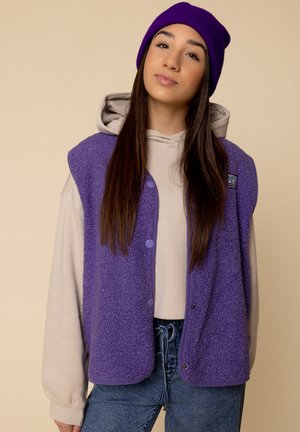 Paars fleece vest met drukknoopsluiting over een beige hoodie, gecombineerd met denim. Kenmerkt zich door een casual snit, gestructureerde stof en effen kleuren.