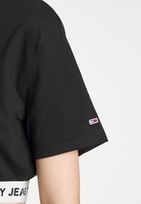 Svart kortärmad skjorta med ett litet rektangulärt rött, vitt och blått logotypmärke nära fållen, bärs av en person med ljus hy.