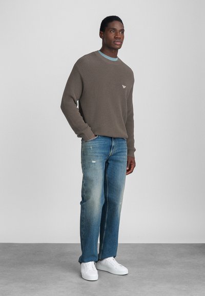 Rjav pulover iz waffle pletenine s okroglo izrezom, v kombinaciji s svetlo modrimi naravenimi ravnimi jeans hlačami in belimi supergami. Preprost logotip na puloverju.