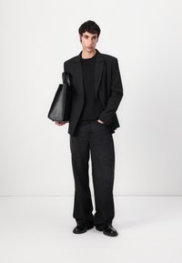 Schwarze Blazer über einem schwarzen Hemd, kombiniert mit dunklen Jeans und schwarzen Schuhen. Hält eine strukturierte schwarze Handtasche. Einfaches, modernes Design.
