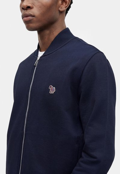 Homme portant une veste zippée bleu marine avec col côtelé et un petit logo cheval multicolore sur la poitrine, sur un fond uni.