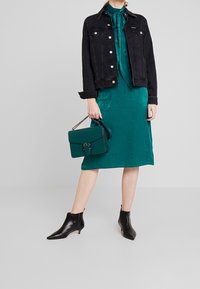 Svart denimjacka över en teal satin klänning, kombinerad med svarta ankelstövlar och en grön handväska med metalldetalj och kedjesnodd.