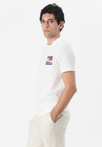 Witte katoenen t-shirt met een kleurrijk grafisch logo op de borst. Korte mouwen en een ronde hals. Gepaard met lichtgekleurde broeken.