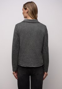 Szara teksturowana kurtka z kołnierzem, długimi rękawami i przednią częścią bez guzików. Połączona z ciemnoszarymi jeansami, pokazana od tyłu.