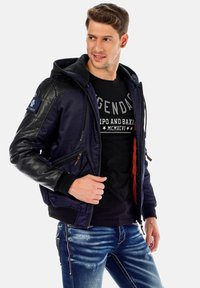 Navyblaue Bomberjacke mit schwarzen Lederärmeln, Reißverschluss-Taschen, elastischen Bündchen und einer abnehmbaren Kapuze. Mit rotem Innenfutter.